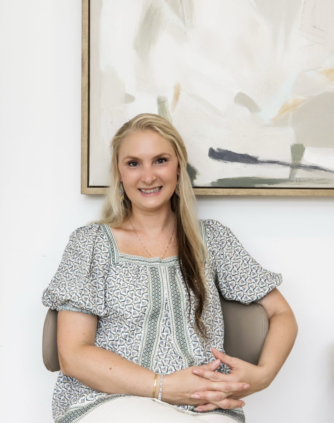 Dr. Shannon, founder of Common Grace Chiropractic & Wellness, seated in the waiting area in a sage green patterned blouse, in front of a large abstract canvas