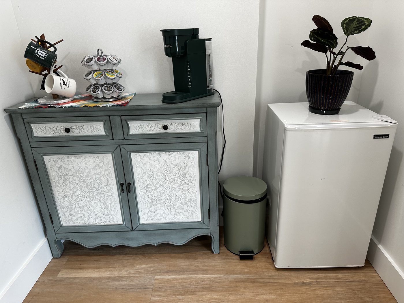 The coffee bar: a sage green cabinet with a coffee machine and mugs on top, a potted plant, and a small fridge alongside