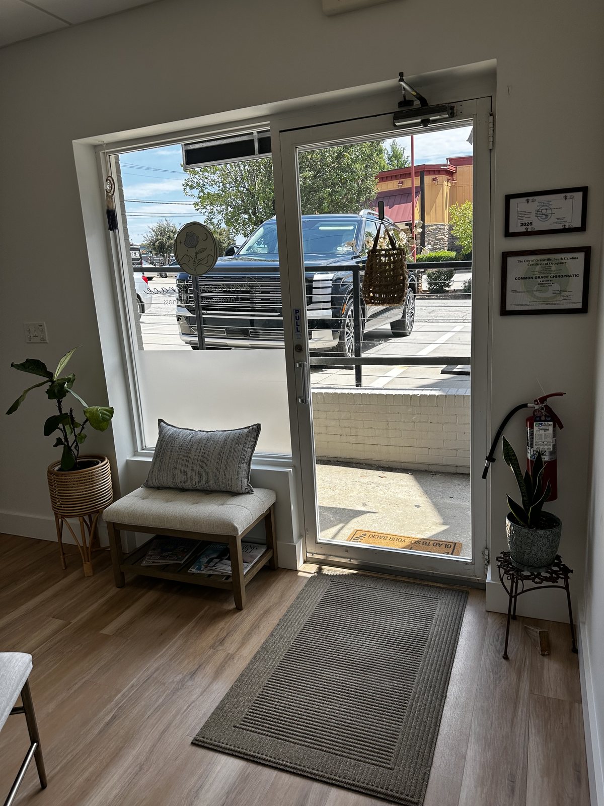 The entrance seen from inside, with daylight coming through the glass door onto a bench, a potted plant and framed certificates on the wall